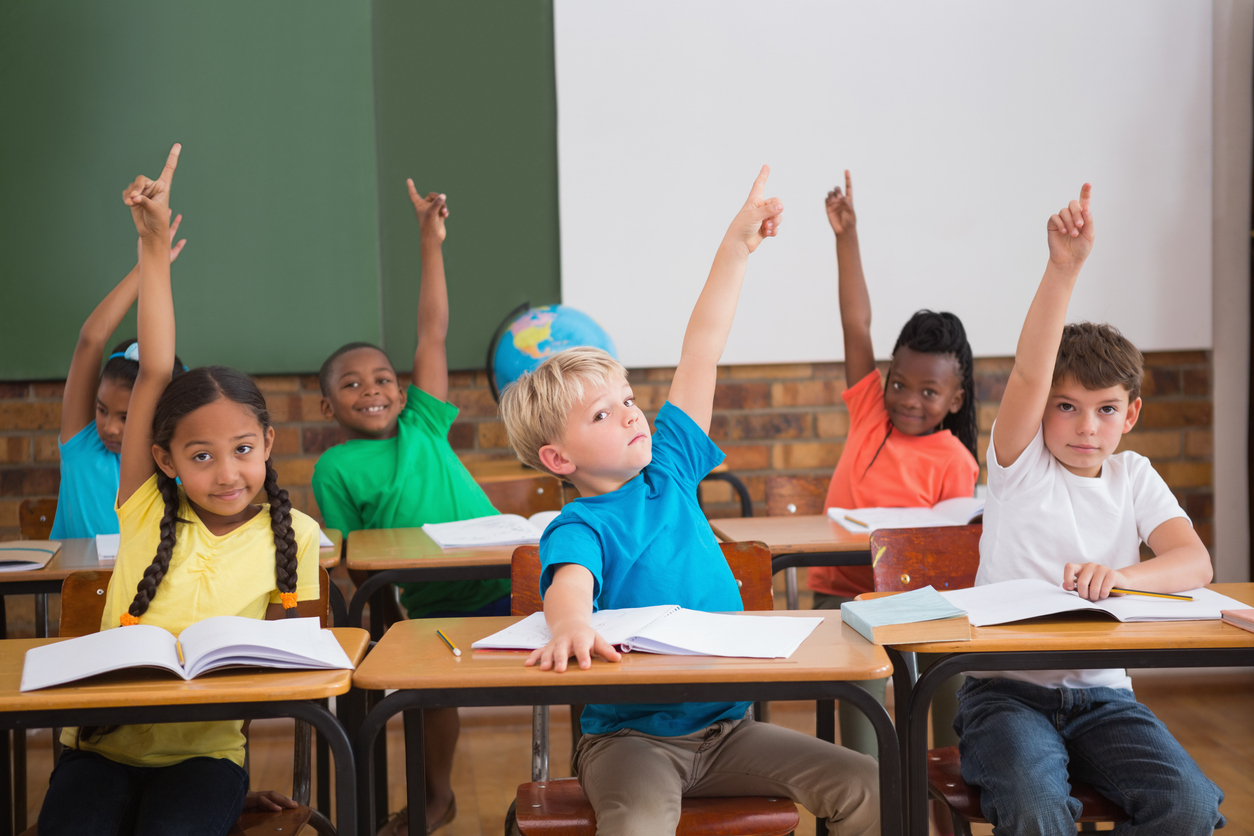 Elementary school students in their desks raising their hands.