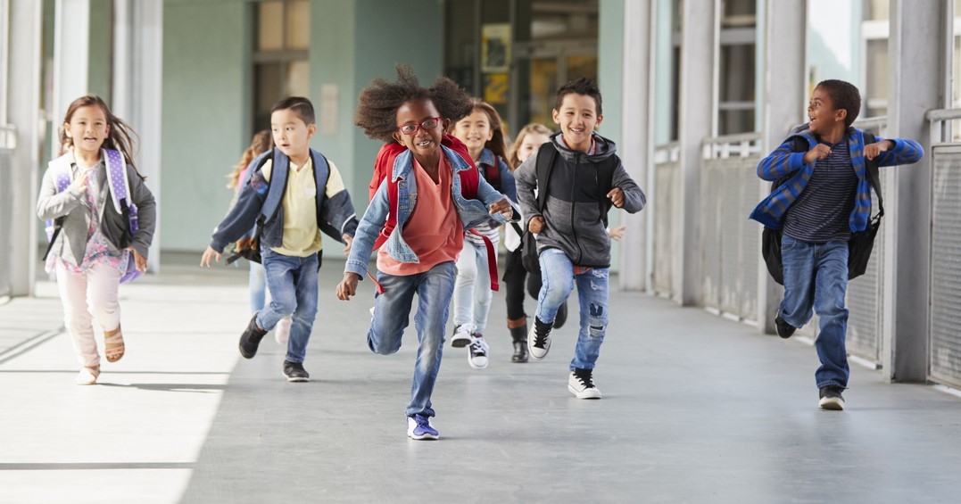 A group of elementary school students wearing backpacks and running in the school hallway.