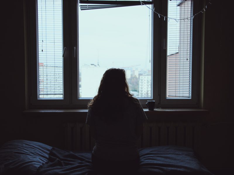 A woman sitting alone in a dark room looking out a window.