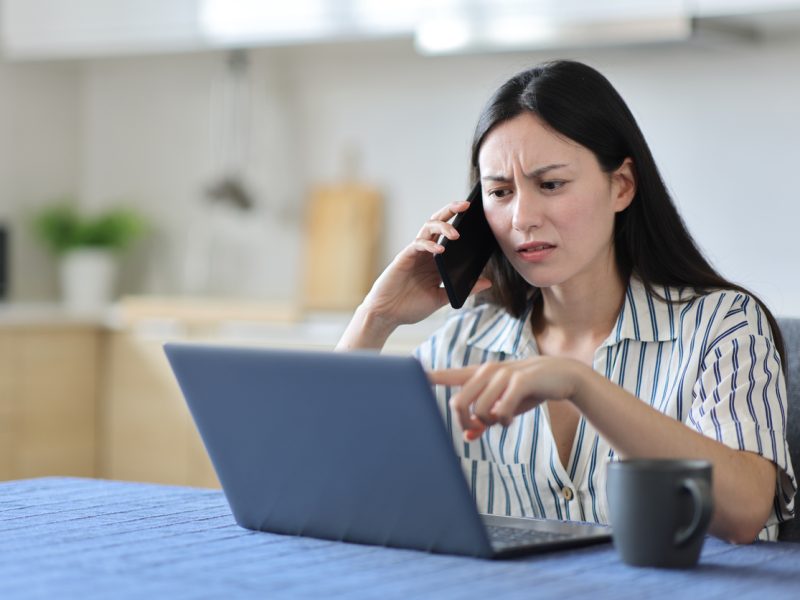 A woman with a cross look on her face talks on her phone while pointing at a laptop screen.